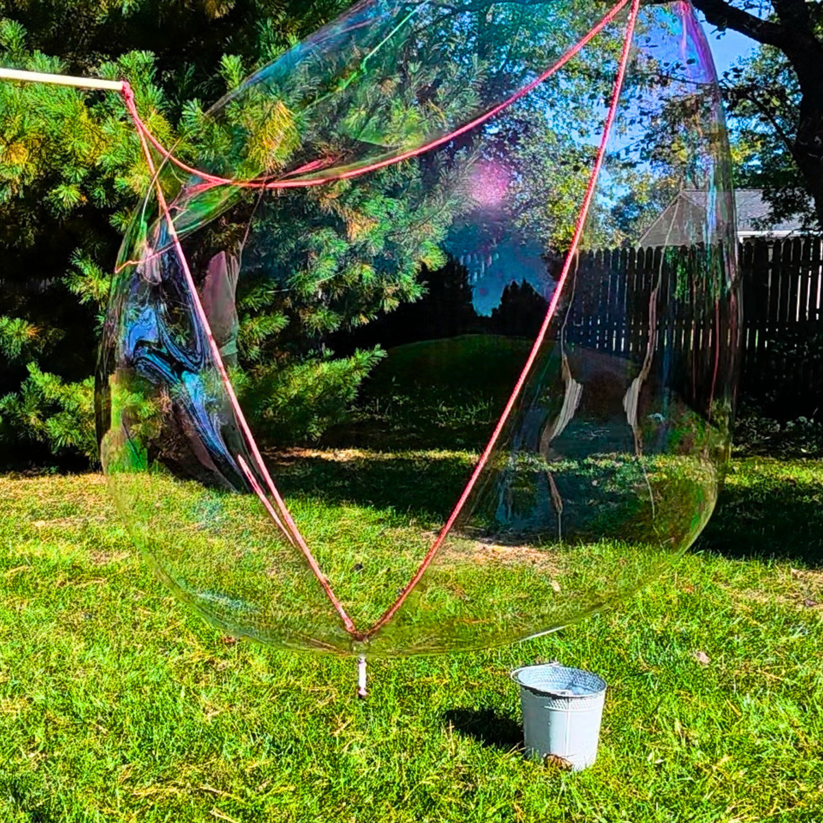 A massive, iridescent giant bubble floating in a sunny backyard next to a decorative bucket, created with One Two Celebrate® Bubble Potion and a handcrafted giant bubble wand
