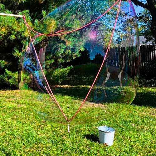 A massive, iridescent giant bubble floating in a sunny backyard next to a decorative bucket, created with One Two Celebrate® Bubble Potion and a handcrafted giant bubble wand