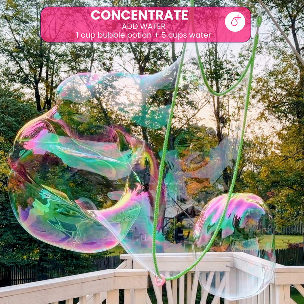 A large, colorful iridescent giant bubble being formed with green rope wands on an outdoor deck, labeled as One Two Celebrate ® Concentrate bubble mix