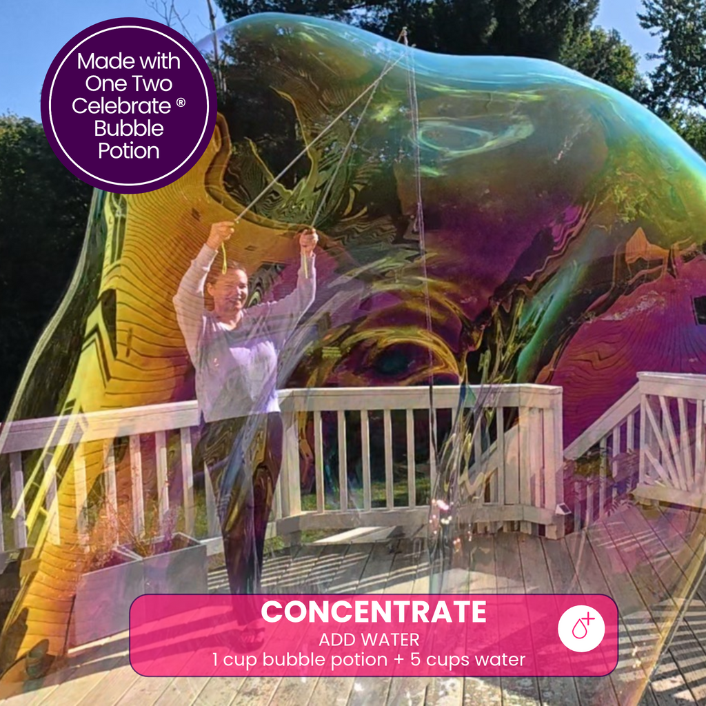 A woman standing inside a massive, iridescent giant bubble on an outdoor deck, featuring One Two Celebrate ® Concentrate bubble potion.