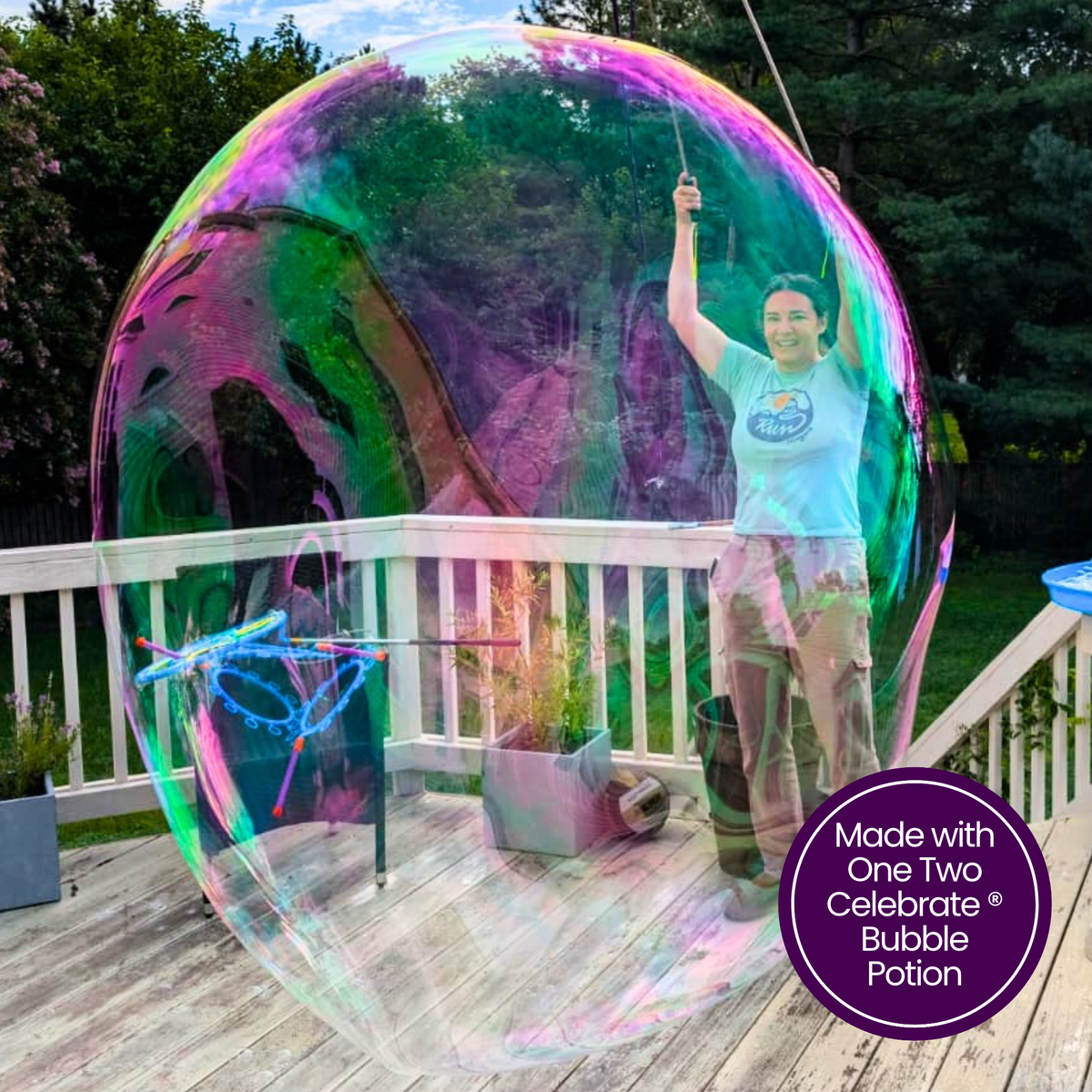 A woman standing inside a massive, vibrant iridescent giant bubble on a wooden deck, holding bubble wands with One, Two, Celebrate® Bubble Potion.